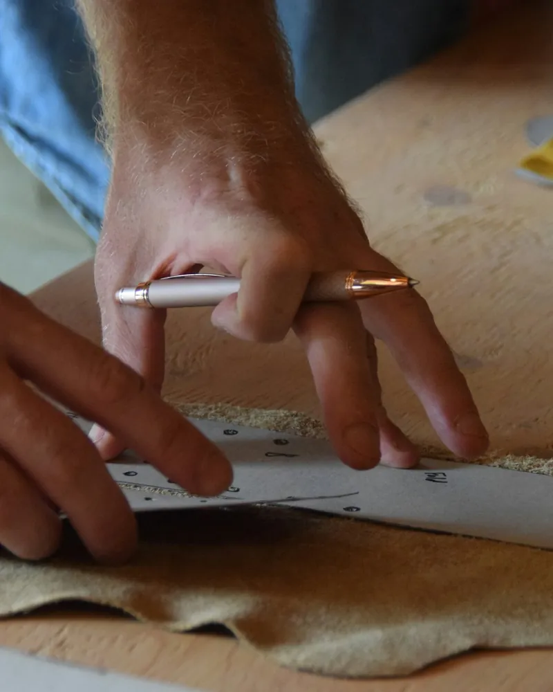 Manos trazando el contorno de un patrón sobre una pieza de cuero natural en un banco de trabajo